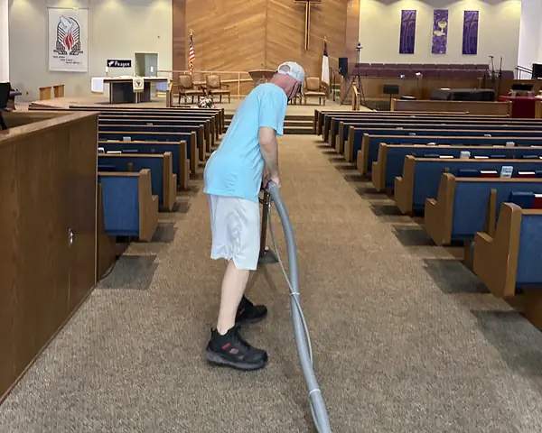 A cleaner in a light blue shirt, a cap and a face mask works a carpet cleaning wand down the centre aisle of a church sanctuary, between rows of blue pews