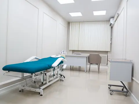 An empty exam room with a teal padded examination table, a desk with two chairs, a wheeled cabinet and a window with vertical blinds