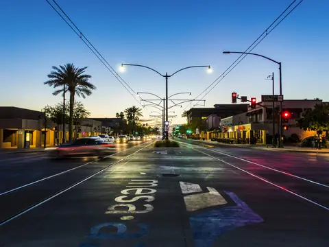 Main Street in downtown Mesa, Arizona at dusk, seen across Center Street, with light rail tracks and overhead wires down the middle, palm trees, lit storefronts and the city's name painted on the pavement