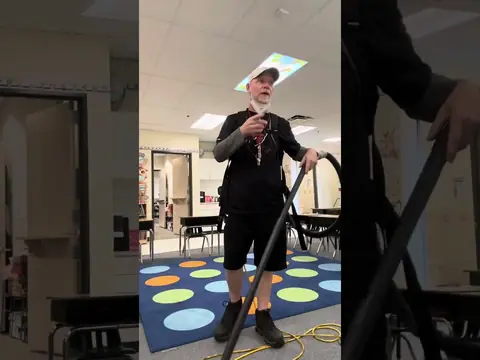 A man in a white cap and a black shirt, wearing a backpack vacuum, holds its wand as he stands on a blue classroom rug with coloured dots