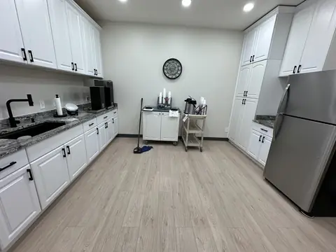 A break room with white cabinets, granite counters, a steel refrigerator and a pale wood-look floor, with a string mop leaning by a coffee cart