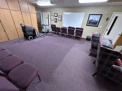 A carpet extractor with its hoses and wand on purple carpet in a meeting room with rows of padded chairs, the cleaned strip of carpet showing darker beside the rest