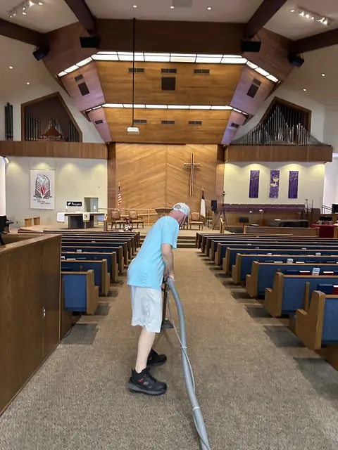A cleaner in a light blue shirt and a cap works a carpet cleaning wand down the aisle of a church sanctuary, with blue pews on both sides and a wooden cross on the far wall