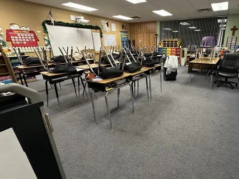 A classroom with chairs turned up on the desks on blue-grey carpet, a whiteboard and a wooden cross on the far wall