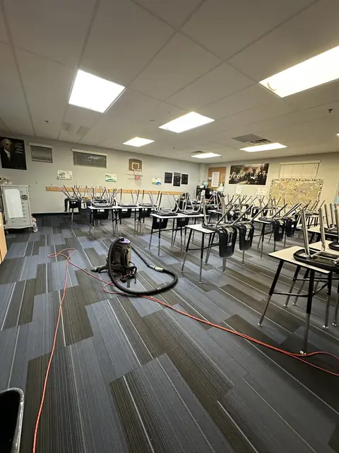 A backpack vacuum and its orange cord on the carpet tiles of a classroom, with the chairs turned up on the desks
