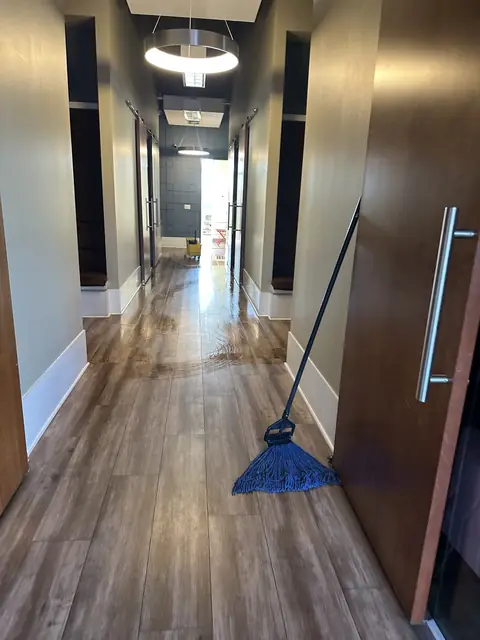 A corridor with a wood-look floor still wet and streaked from mopping, a blue string mop leaning on a sliding door in the foreground and a yellow mop bucket at the far end