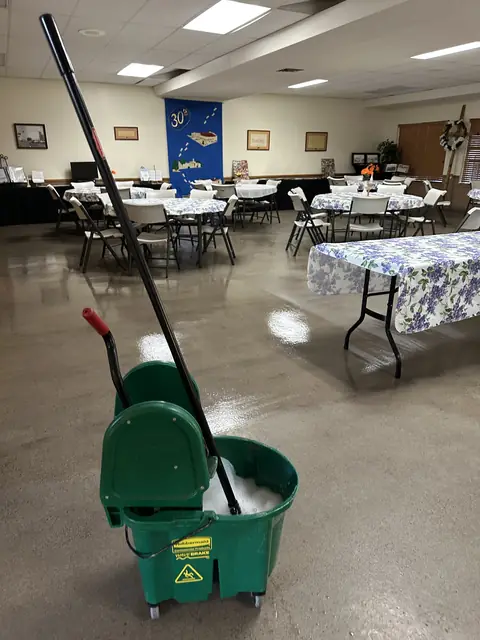 A green mop bucket full of suds on the wet, sealed floor of a hall set with tables under flowered cloths