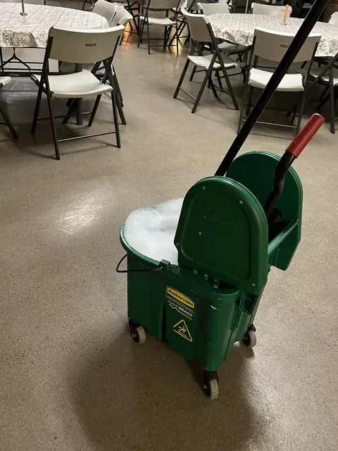 A green mop bucket full of suds on the sealed floor of a hall with round tables under lace cloths and white folding chairs