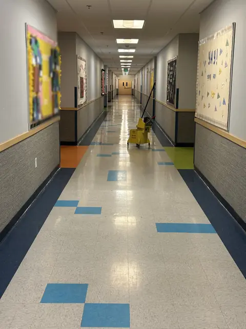 A long school hallway with a glossy tile floor set with blue tiles, bulletin boards on both walls and a yellow mop bucket at the far end
