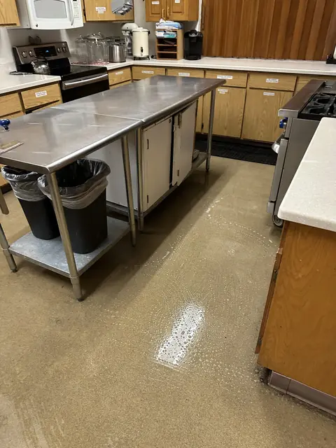 A kitchen floor wet with suds beside a steel work table, with wooden cabinets, a stove and two lined trash bins