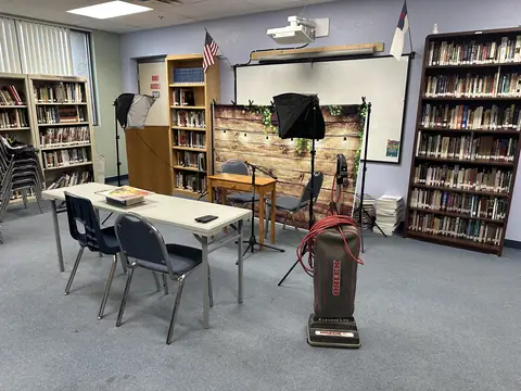 An upright vacuum with its red cord on blue carpet in a library room with full bookshelves, a folding table and a small recording set