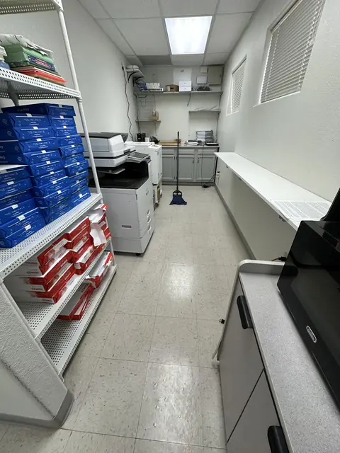 A mail and copy room with shelves of blue and red mail trays, two copiers and a speckled tile floor, with a broom leaning at the far end