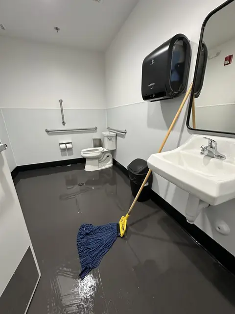 An accessible restroom with a toilet and grab bars, a wall sink, a paper towel dispenser and a mirror, its dark floor wet and shining behind a string mop