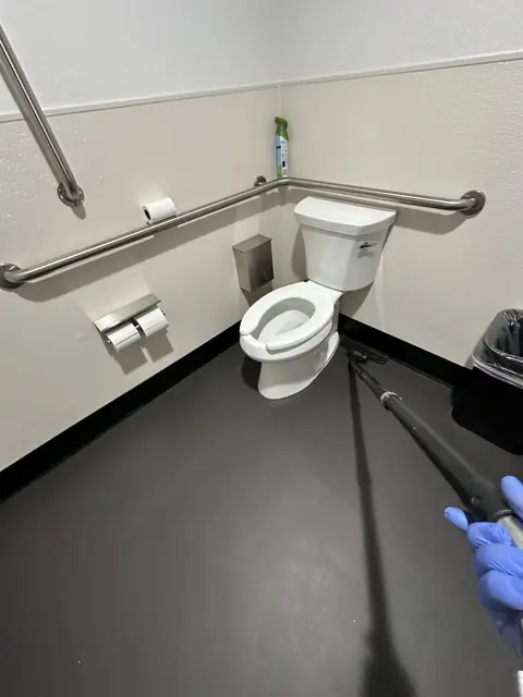 A gloved hand holding a mop handle beside a toilet with grab bars, on the dark floor of an accessible restroom