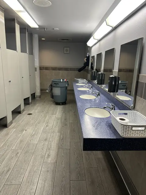 A restroom with a long blue counter of four sinks under a row of mirrors, a line of stalls and a grey bin at the far end