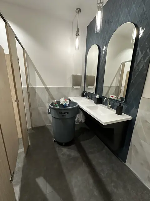 A restroom with two arched mirrors on a dark blue tiled wall, a white floating counter with two sinks, pendant lamps and a dark tile floor, with a grey bin of spray bottles and cloths beside the counter