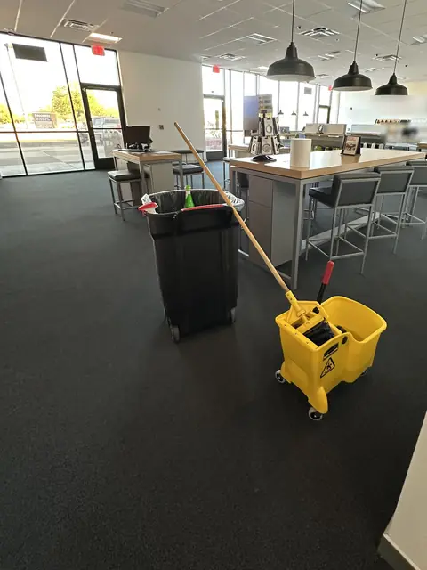A yellow mop bucket and a black bin on the dark carpet of a store, with display tables, stools and pendant lamps behind