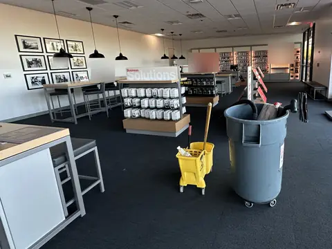 A yellow mop bucket and a grey bin holding a backpack vacuum on the dark carpet of a phone store, with display tables, shelves of accessories and pendant lamps