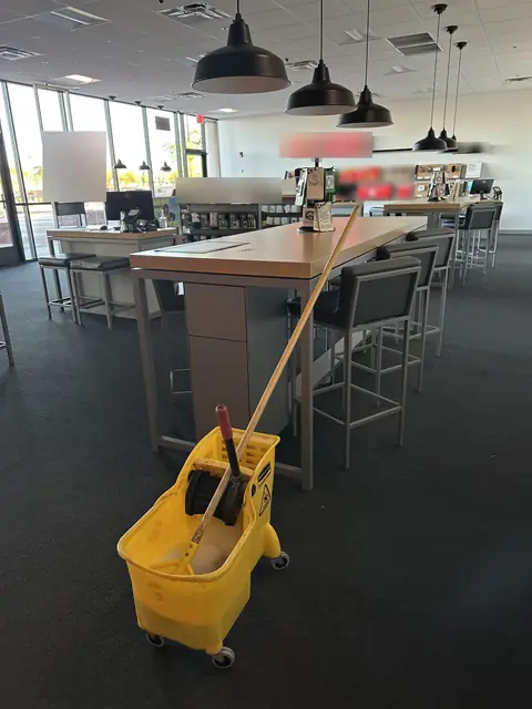A yellow mop bucket on the dark carpet of a store beside a long display table with stools, under black pendant lamps