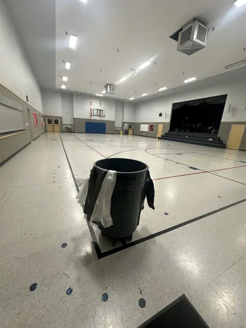A school gym and stage with a pale tile floor marked with court lines, and a grey bin hung with liners in the foreground