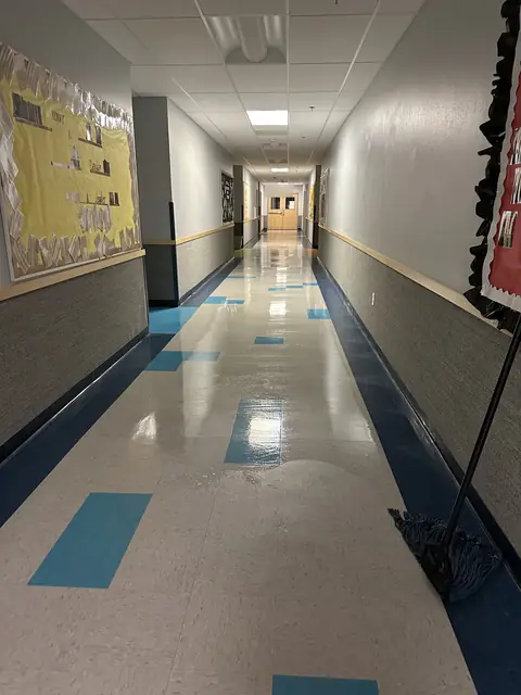 A school hallway floor wet and shining along its length, with a string mop leaning on the wall in the foreground