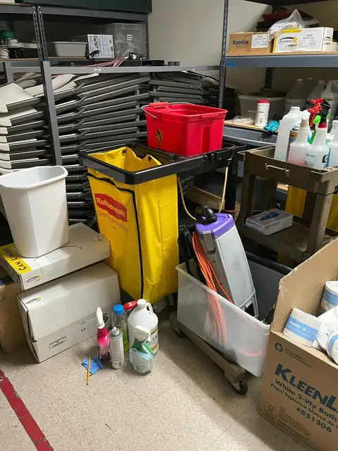 A supply room with a janitor's cart and its yellow bag, a red caddy, spray bottles, a vacuum and boxes of liners on steel shelves