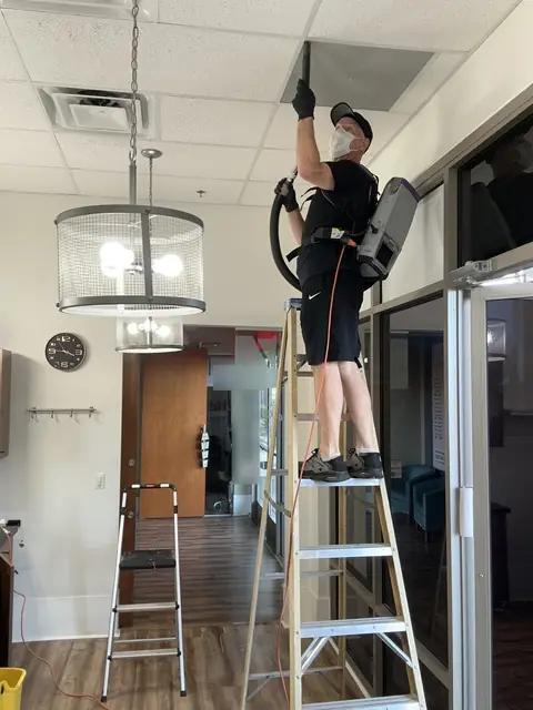 A crew member in black, wearing a cap, a face mask and a backpack vacuum, stands on a ladder and holds the vacuum wand to a ceiling vent